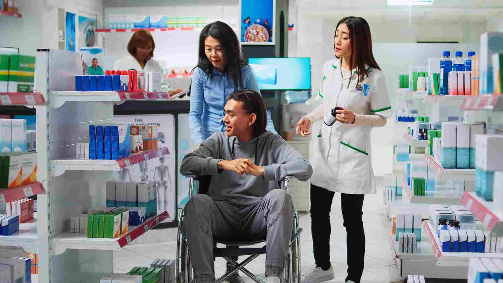 Pharmacist assisting a wheelchair-bound patient and caregiver in a modern hospital pharmacy, representing inclusive and efficient patient services enabled by HMS.