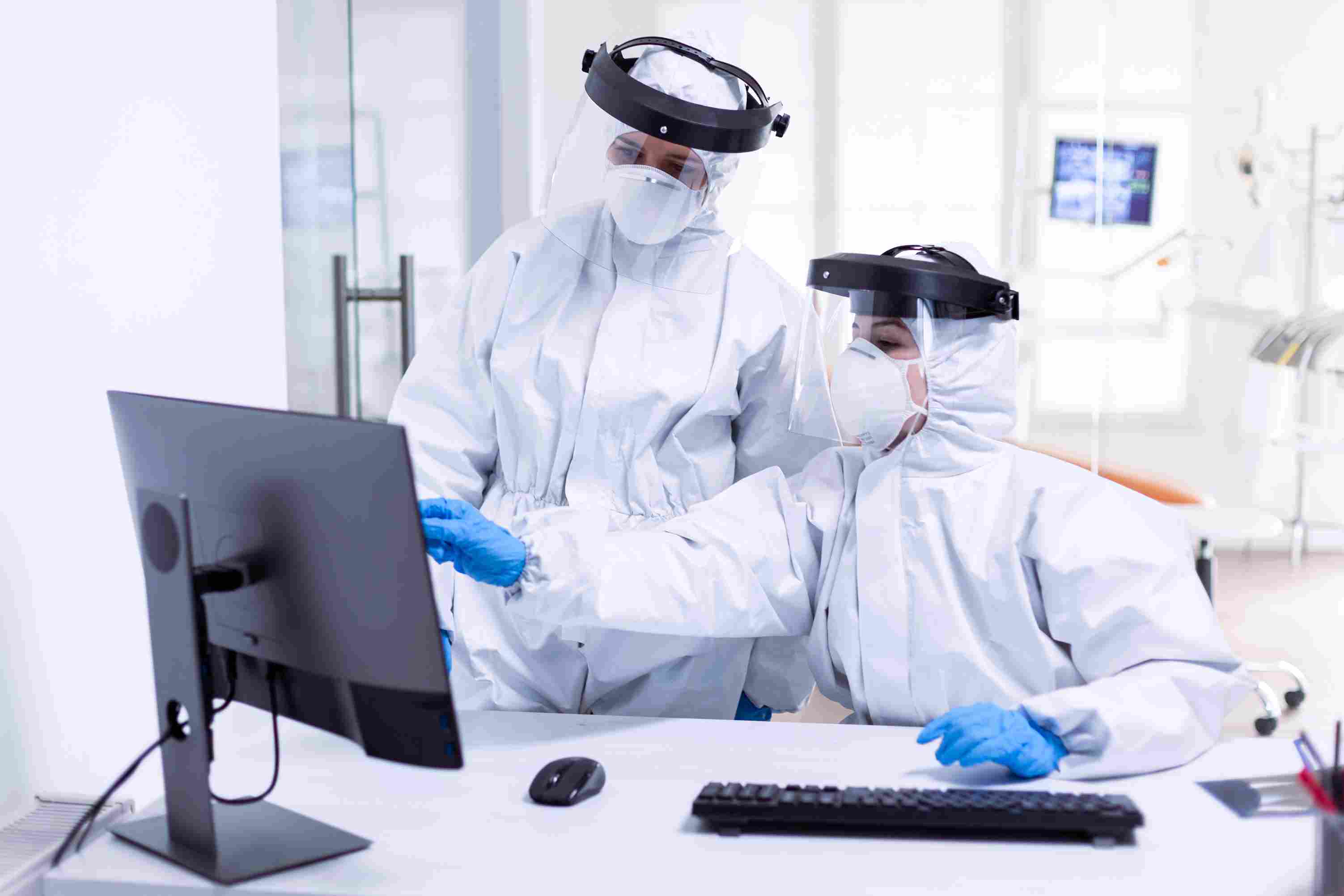 Two healthcare workers in full protective gear collaborate at a computer workstation, representing the adoption of the QHIE-Hub Platform, Health Information Exchange, and ePrescription & Pharmacy Network for secure, integrated, and patient-centric care in Qatar