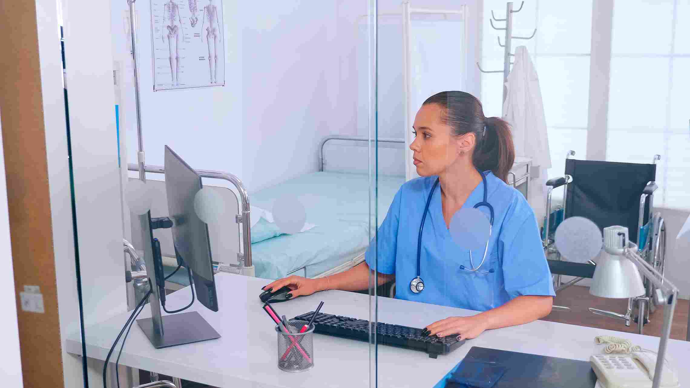 Nurse in scrubs using hospital management software at a computer to handle billing, insurance claims, and patient information