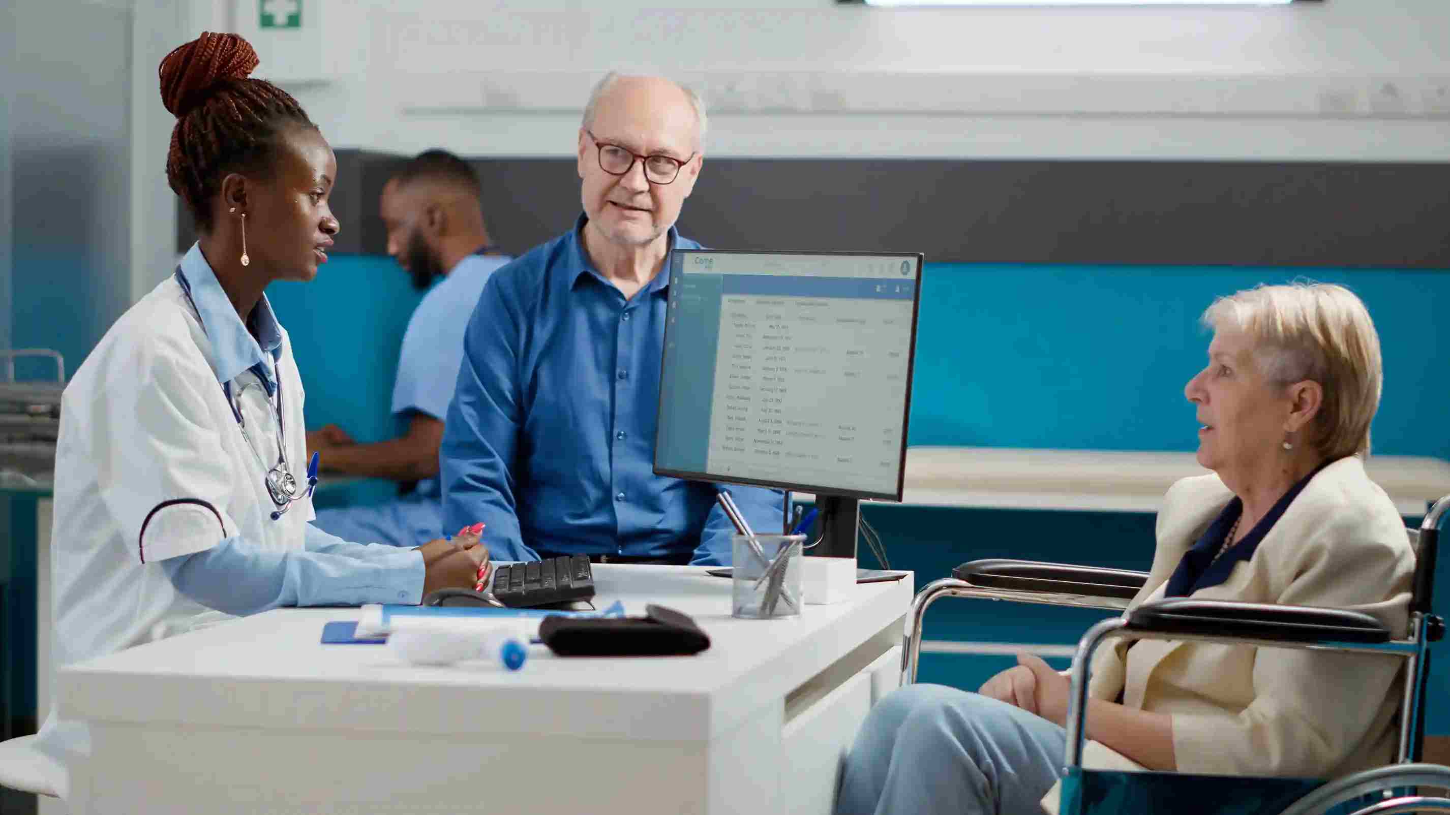 A general practitioner speaks with an elderly woman in a wheelchair and her companion while viewing medical details on a desktop screen, illustrating bedside use of a Patient Management System