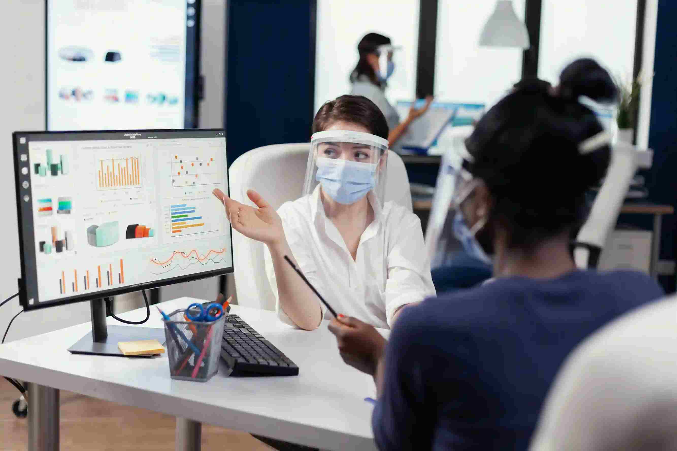 Masked staff review colorful graphs on a wide monitor in an office, representing how a Patient Management System provides real-time operational and financial insights