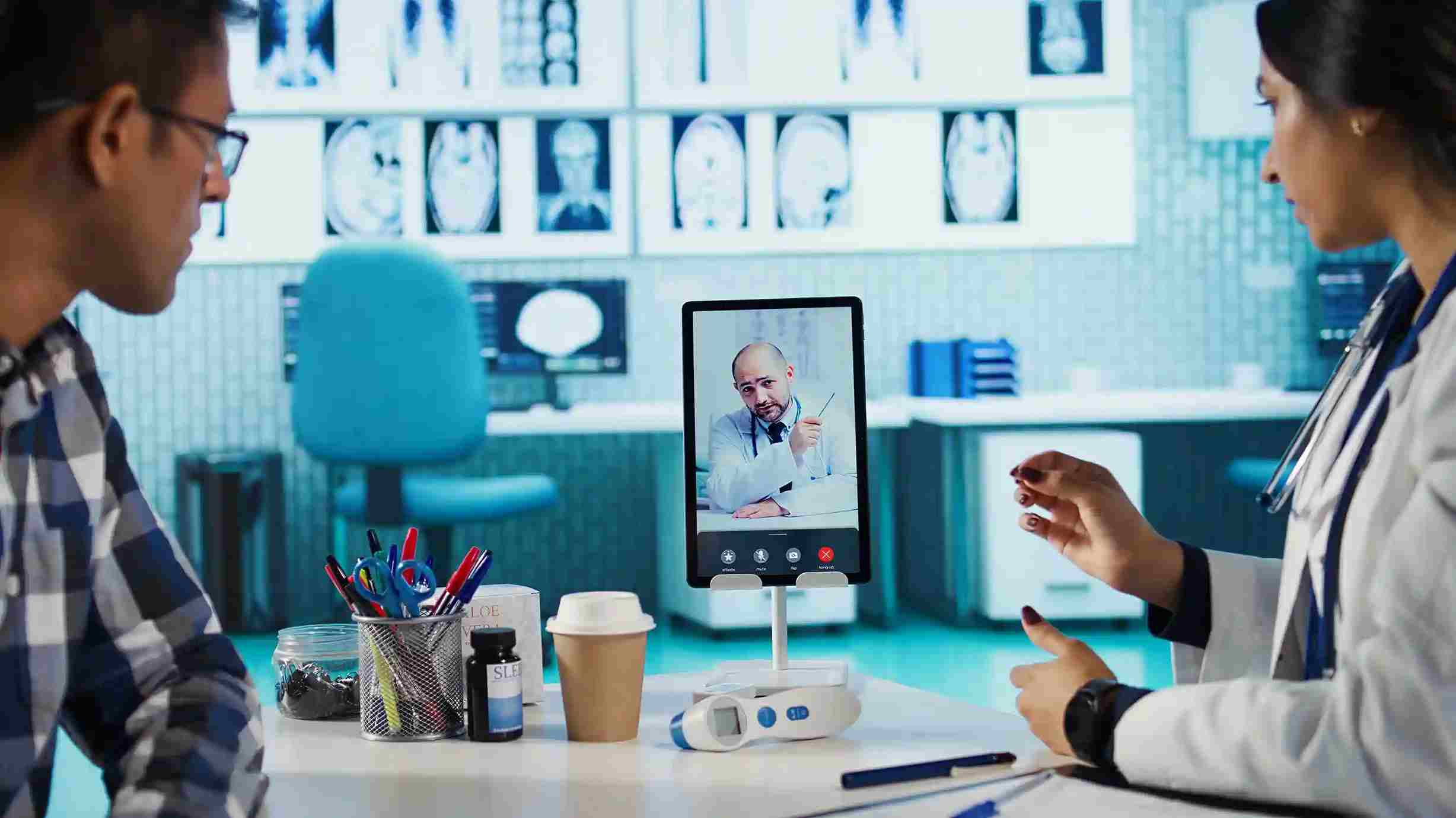 Doctor and patient sit at a desk while another physician appears on a tablet video call, showing how telehealth connects into a Patient Management System for coordinated care