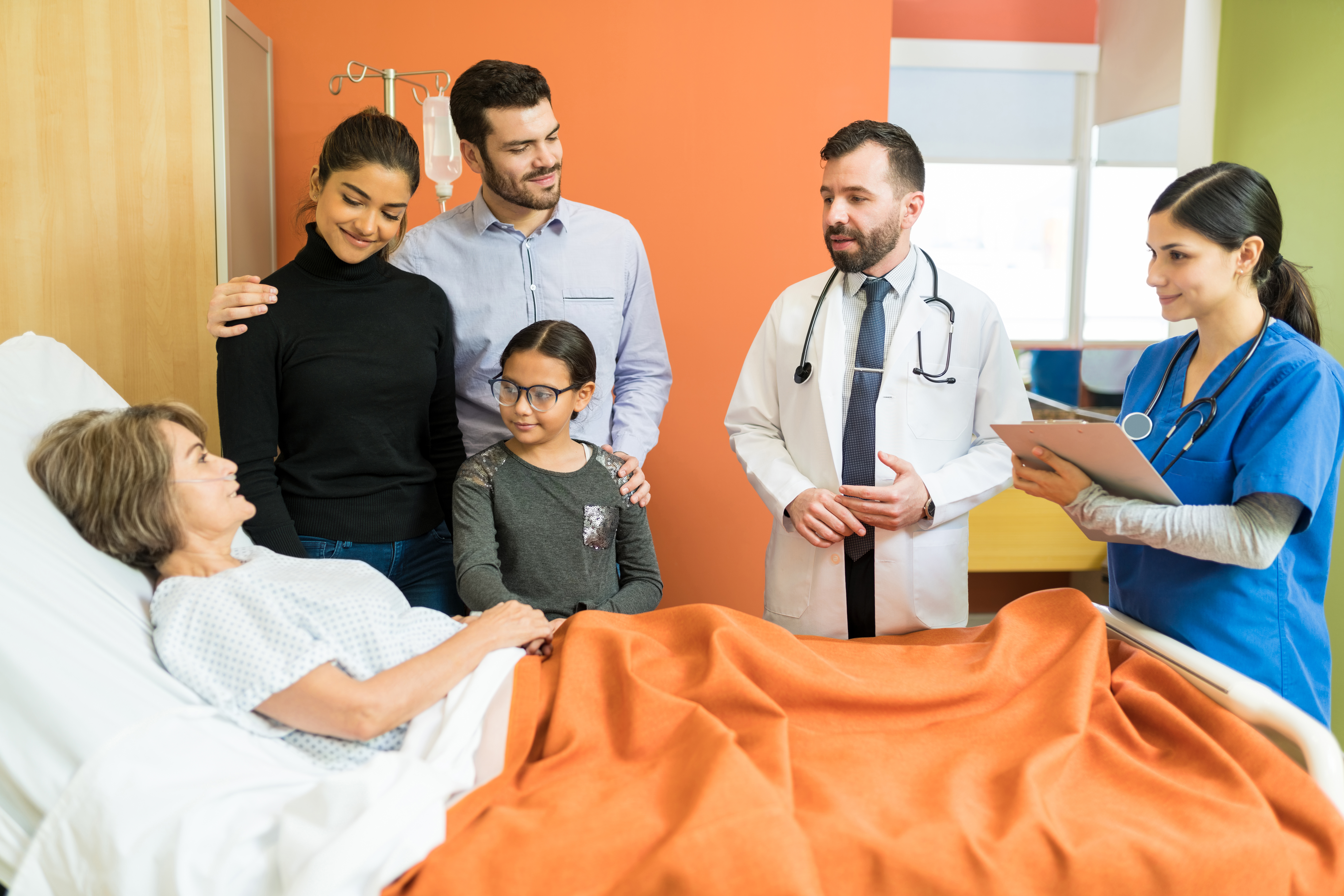 Doctors and nurses discuss treatment with a patient and family at the bedside, showing coordinated inpatient workflows that help reduce delays and improve patient flow