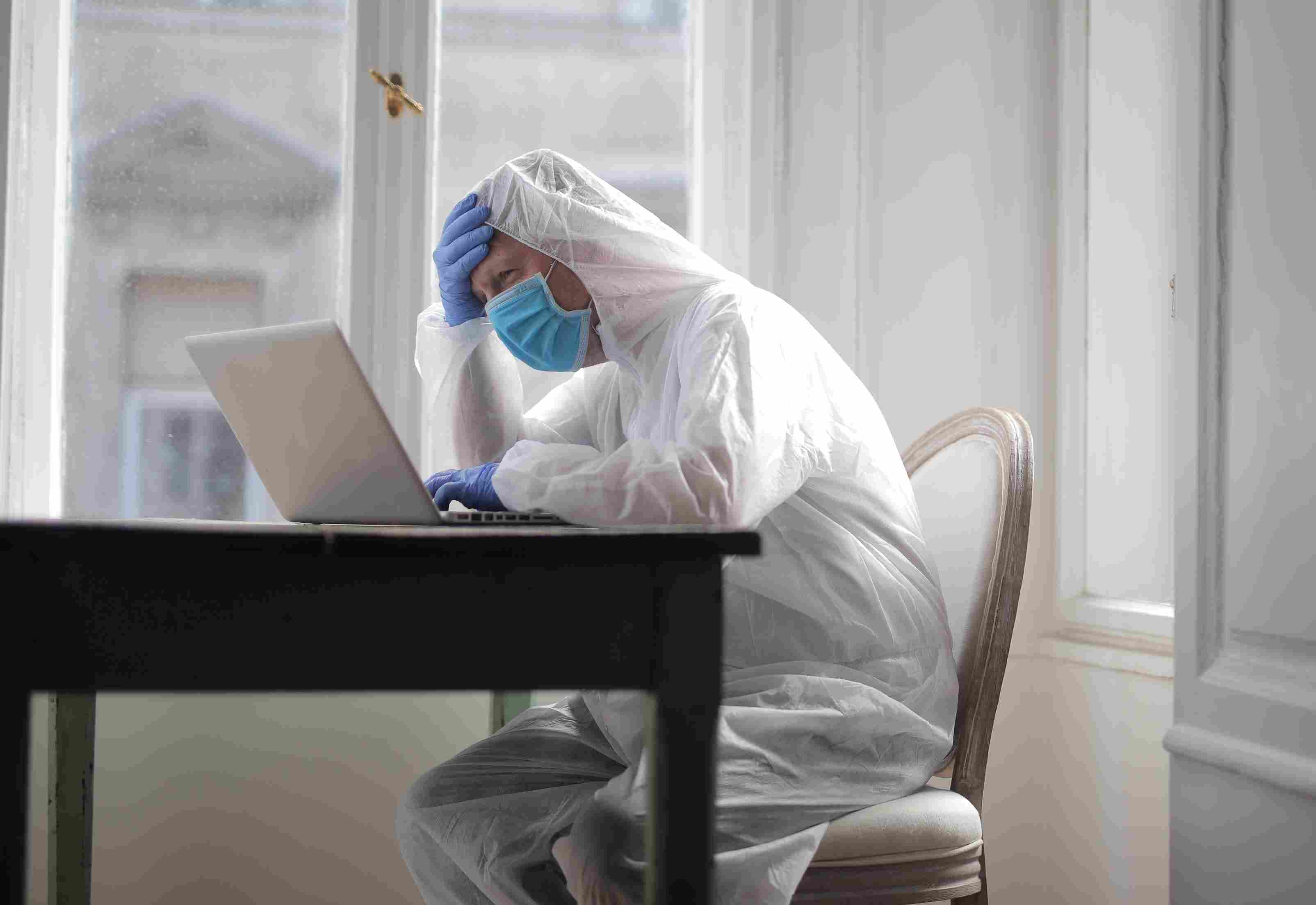 Healthcare worker in protective equipment showing exhaustion while managing administrative tasks on laptop