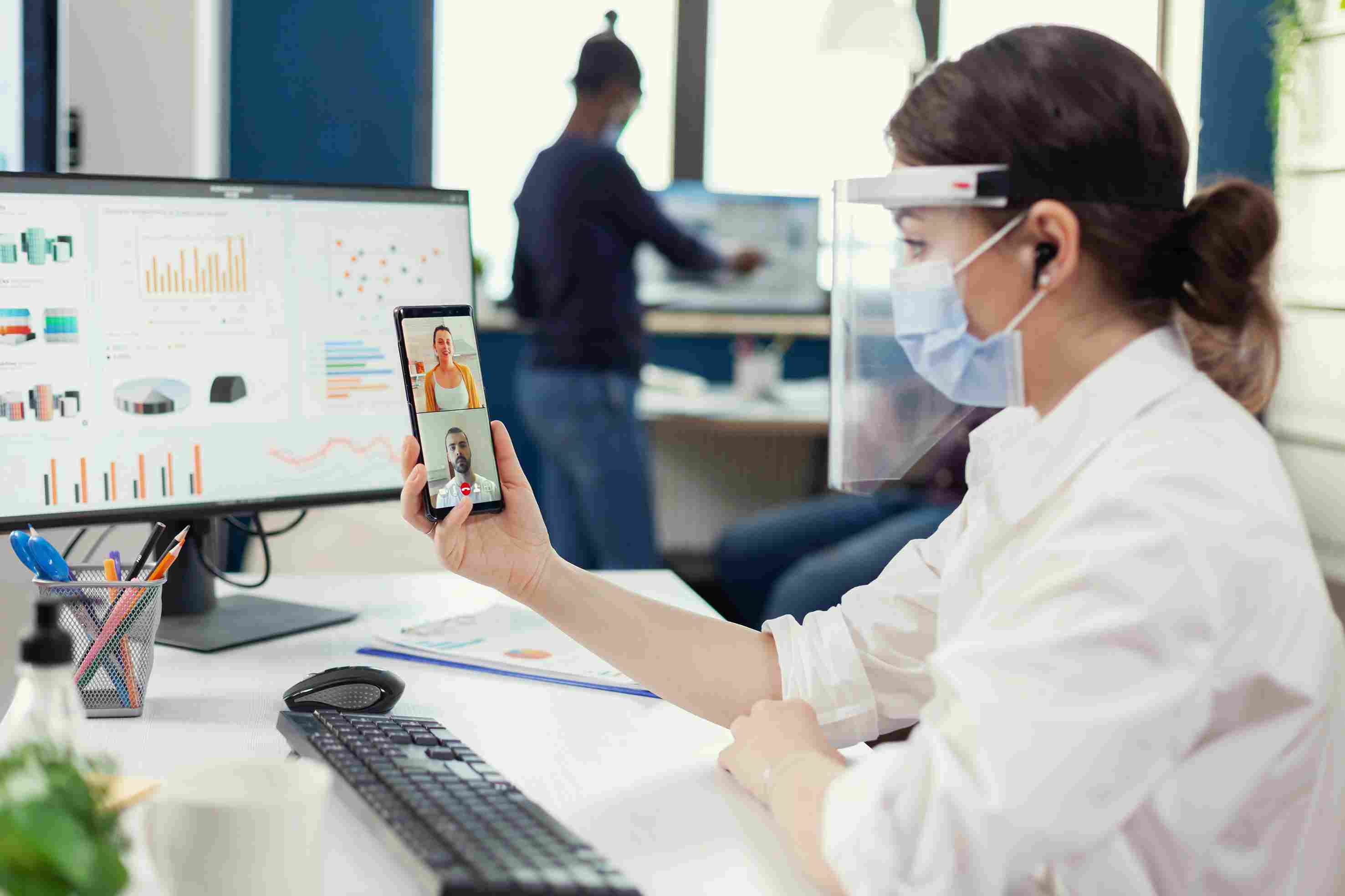 A masked healthcare professional in a face shield holds a smartphone video call while multi-location performance dashboards display on a monitor behind her, highlighting remote clinic management and analytics capabilities.