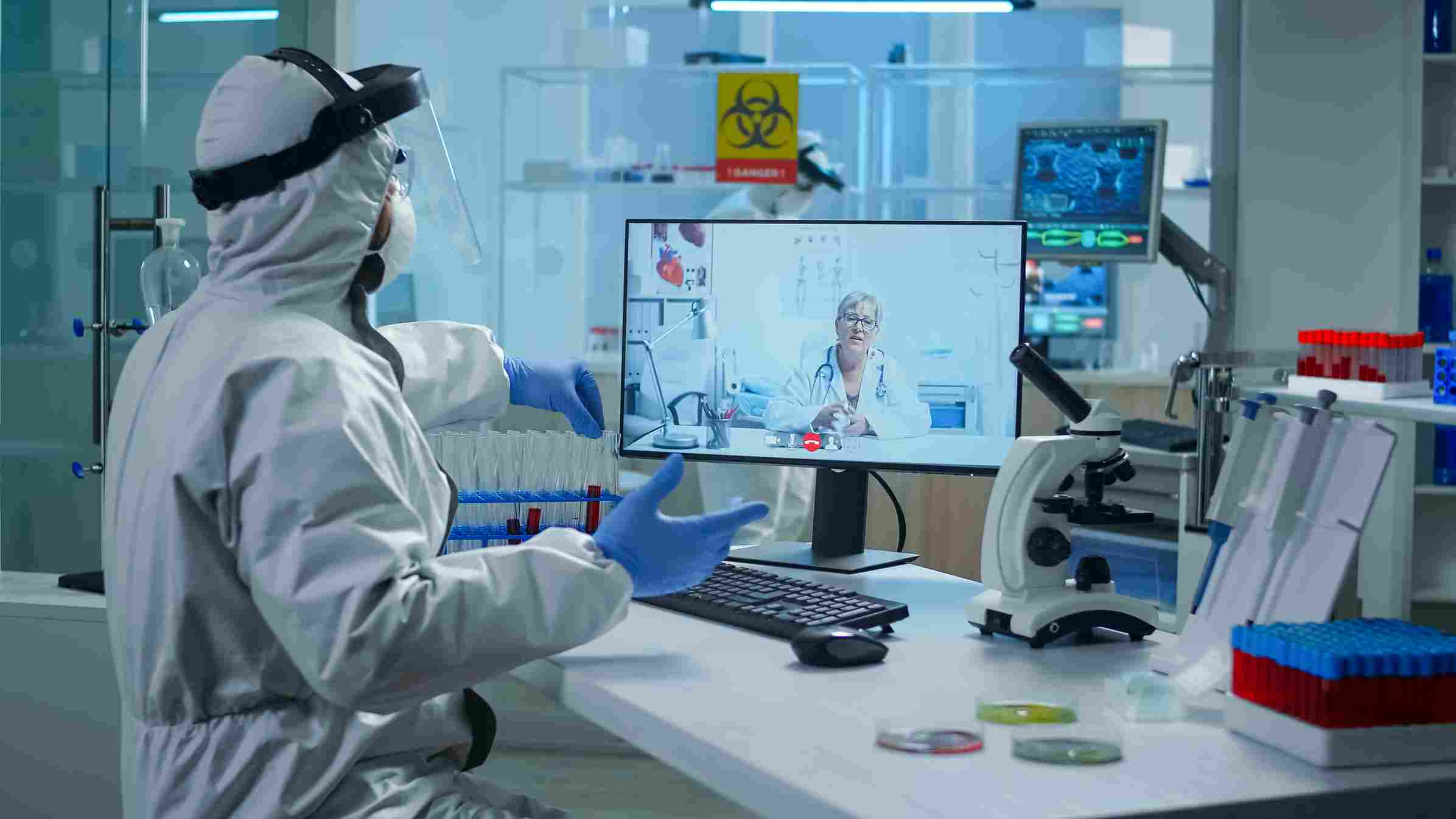 Lab technician in protective equipment consulting senior doctor via video call using Laboratory Management Software for remote collaboration