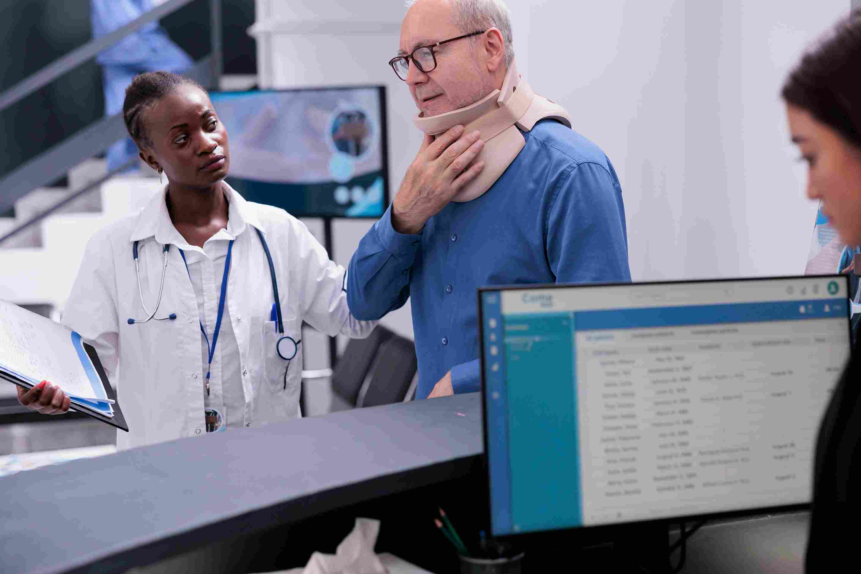 A physician in a white coat attends to an elderly patient wearing a cervical collar at a hospital reception desk, with a patient appointment management system visible on the receptionist's monitor in the foreground.