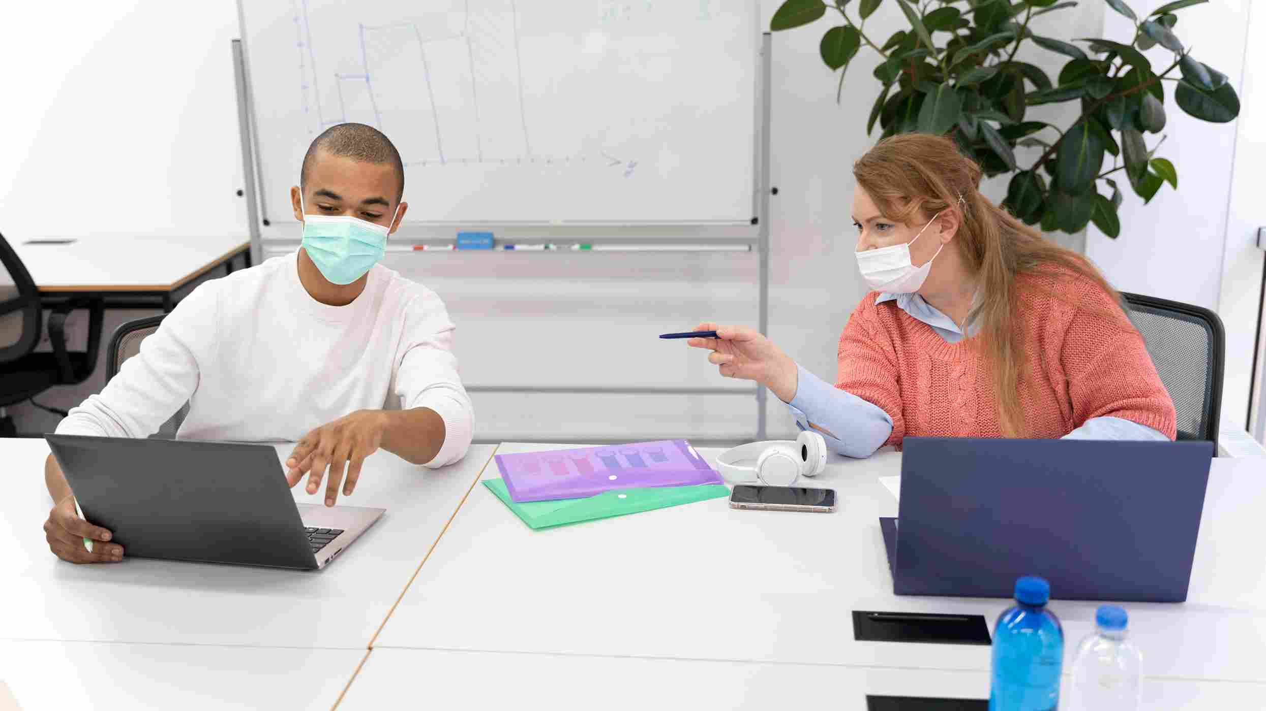 Two healthcare office employees wearing face masks collaborate over laptops at a shared workspace, representing coordinated multi-location clinic scheduling and staff communication.