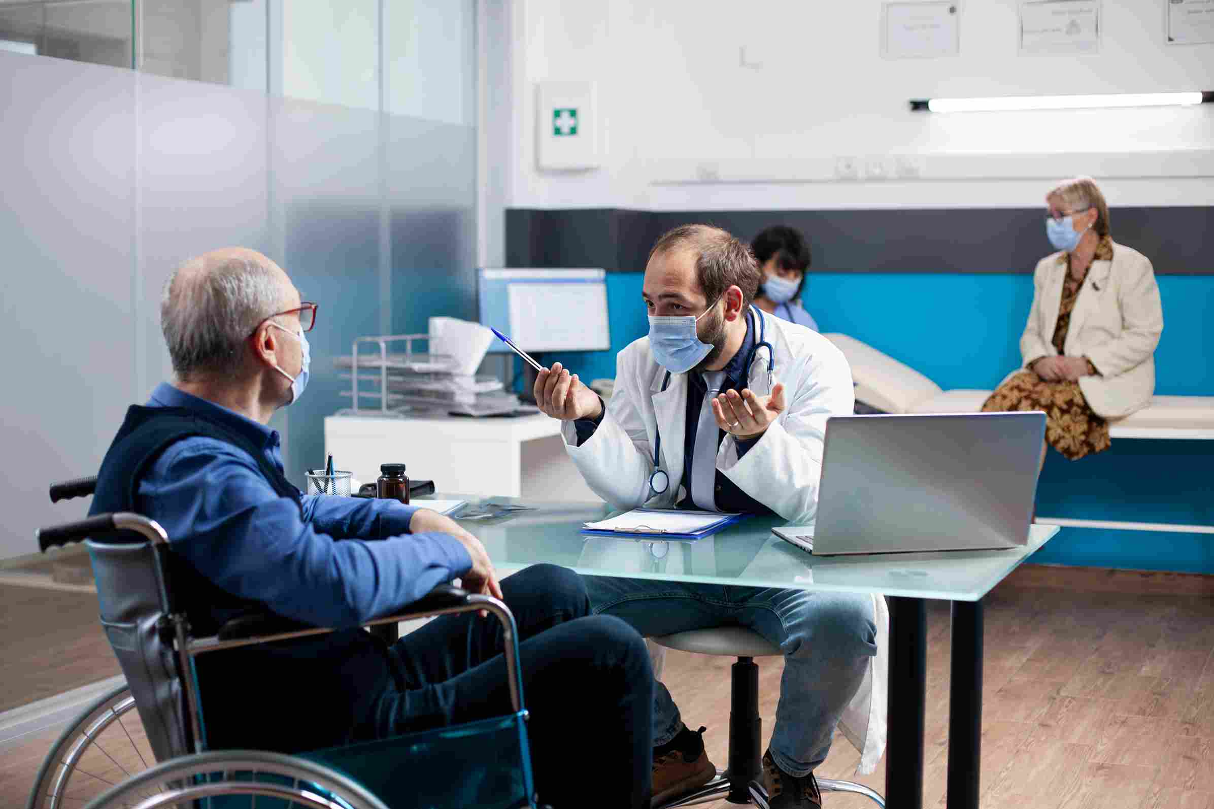 A doctor having a focused consultation with an elderly patient seated in a wheelchair in a hospital setting with clinical staff visible in the background, representing the Patient Rights and Education and Access Assessment and Continuity of Care chapters of NABH 6th Edition accreditation that require documented patient consent, education, and coordinated care plans supported by a Hospital Management System