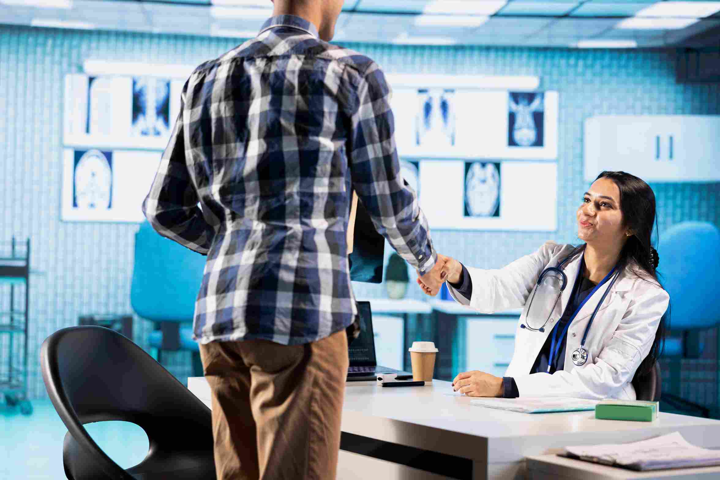 A smiling female doctor in a white coat extending a handshake to a patient in a clinical room with medical imaging scans displayed on the wall behind her, representing the improved patient trust and care experience that results when a hospital selects an HMS vendor with reliable support, transparent data policies, and genuine post go-live commitment