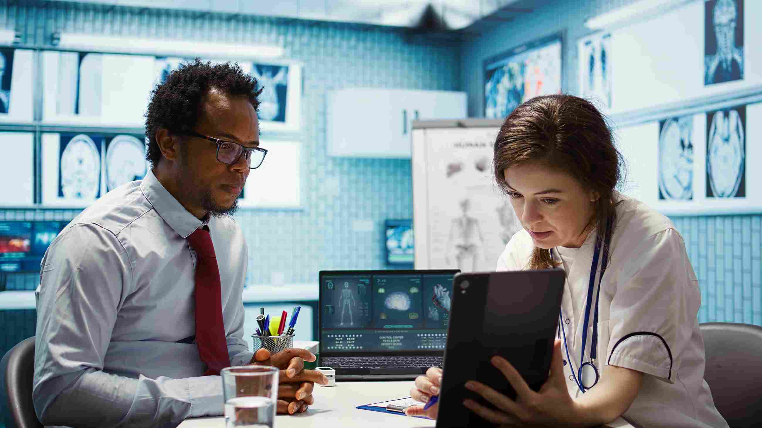 A hospital administrator in formal attire and a doctor in a white coat reviewing medical imaging scans and HMS performance data on a laptop and tablet in a clinical setting surrounded by brain scans, representing the financial and operational data analysis that helps hospital leaders measure the return on investment of their Hospital Management System
