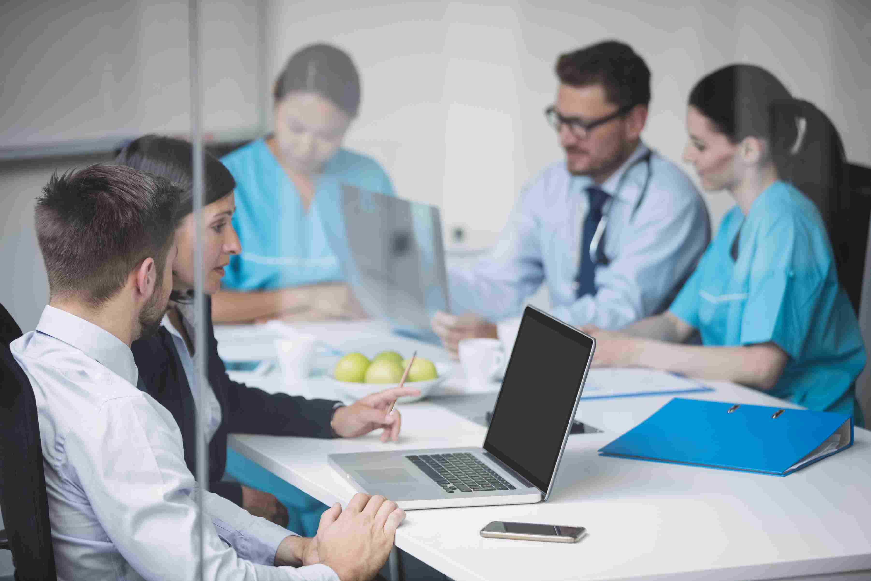 A mixed team of hospital administrators and medical professionals in scrubs gathered around a conference table with a laptop and medical files, representing the discovery and scoping phase of a Hospital Management System implementation