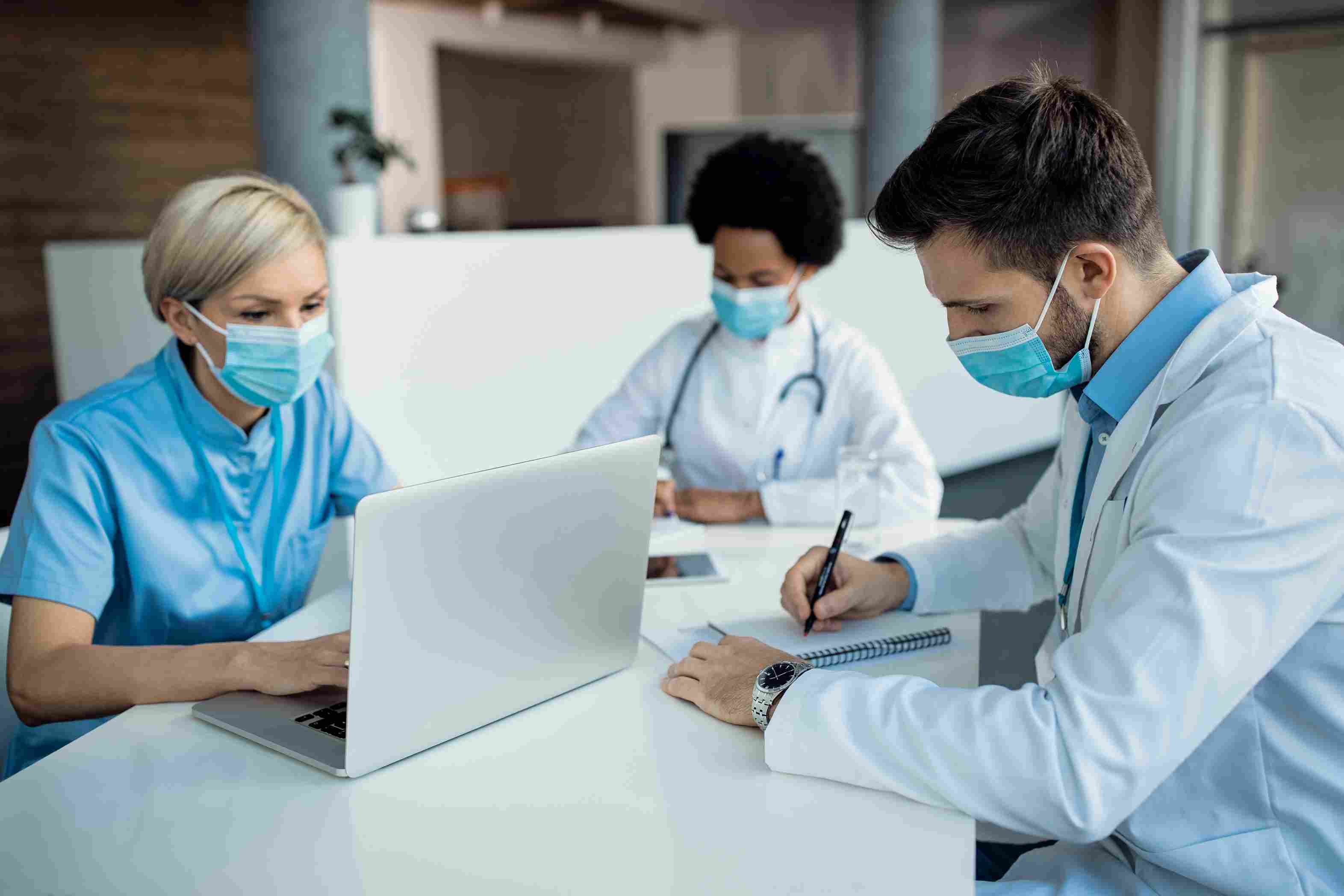A nurse in blue scrubs working on a laptop alongside a doctor in a white coat taking written notes while a colleague reviews documents in the background, representing the day to day clinical team preparation steps required to achieve Milestone 1 and Milestone 2 ABDM compliance in Indian hospitals in 2026