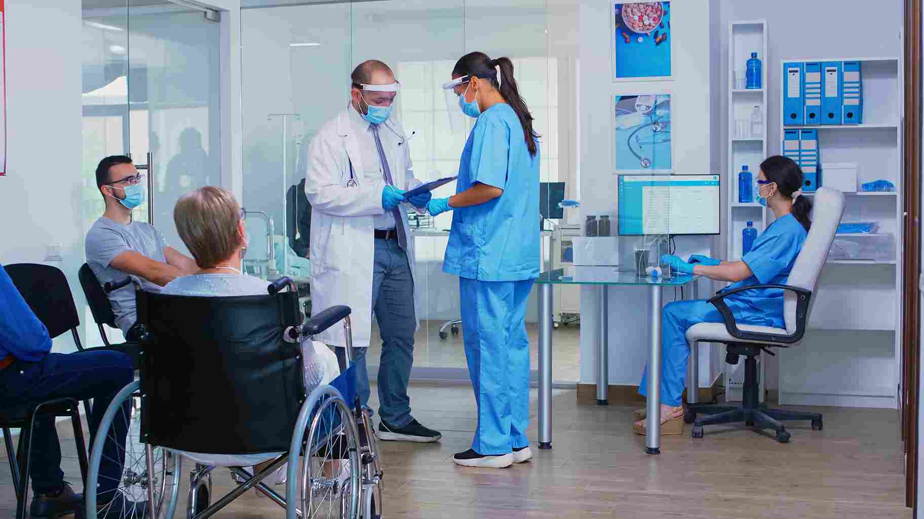 A doctor and nurse reviewing patient documentation on a clipboard at a hospital reception desk while patients wait in the background, illustrating the administrative workflows that a Hospital Management System helps coordinate