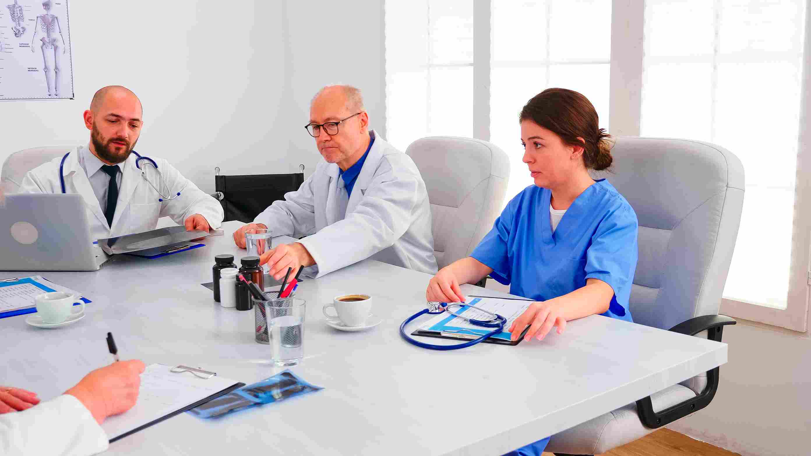 A group of doctors and clinical staff in white coats gathered around a conference table reviewing radiography reports and clinical documentation, representing the Continuous Quality Improvement and Information Management System chapters of NABH 6th Edition accreditation where hospital teams must demonstrate quality indicator tracking, incident reporting, and complete audit trails through integrated Hospital Management System modules