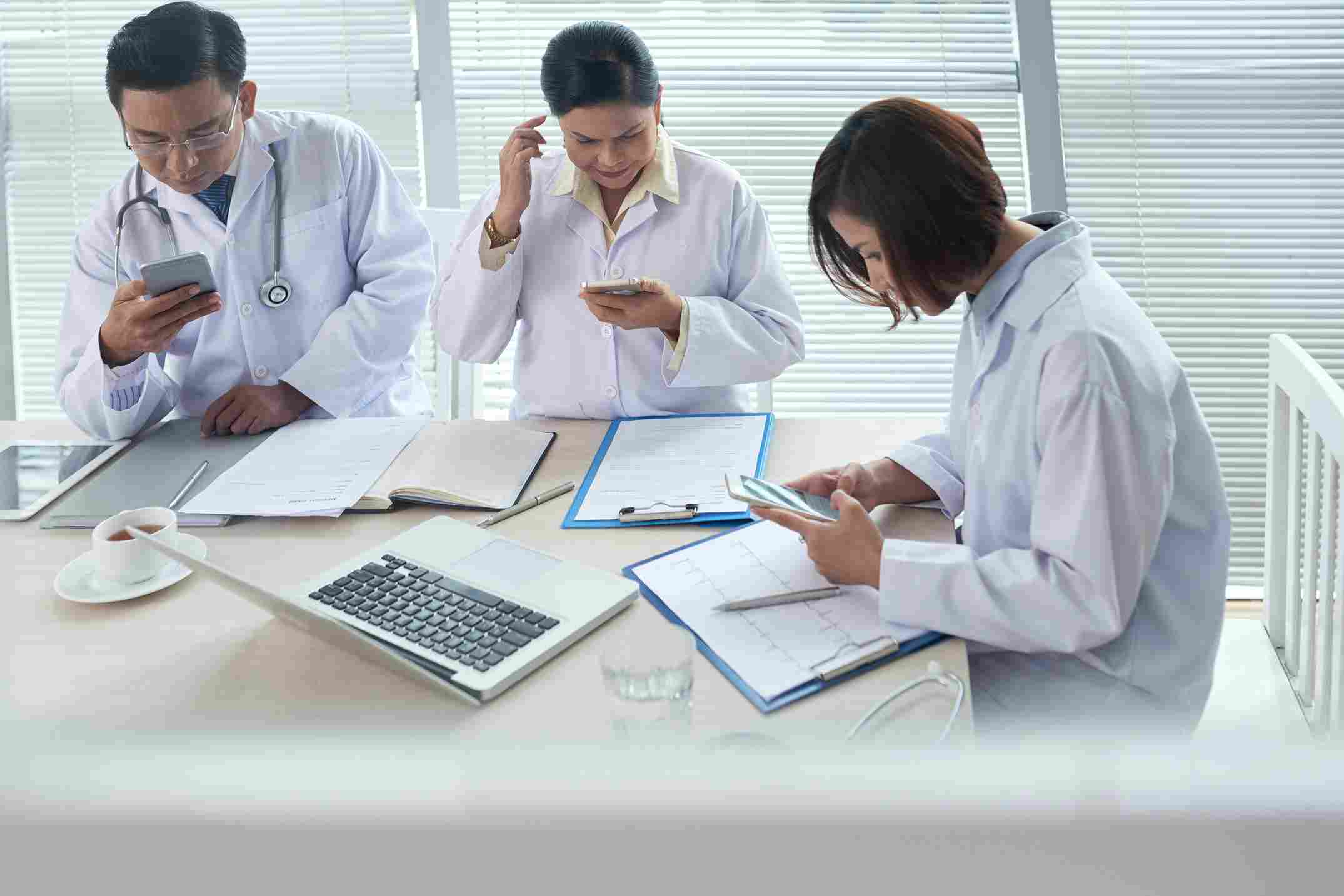 Three doctors in white lab coats using smartphones and reviewing paper reports and clipboards at a desk with a laptop, illustrating the administrative burden on clinical staff that HMS software reduces to improve productivity and ROI