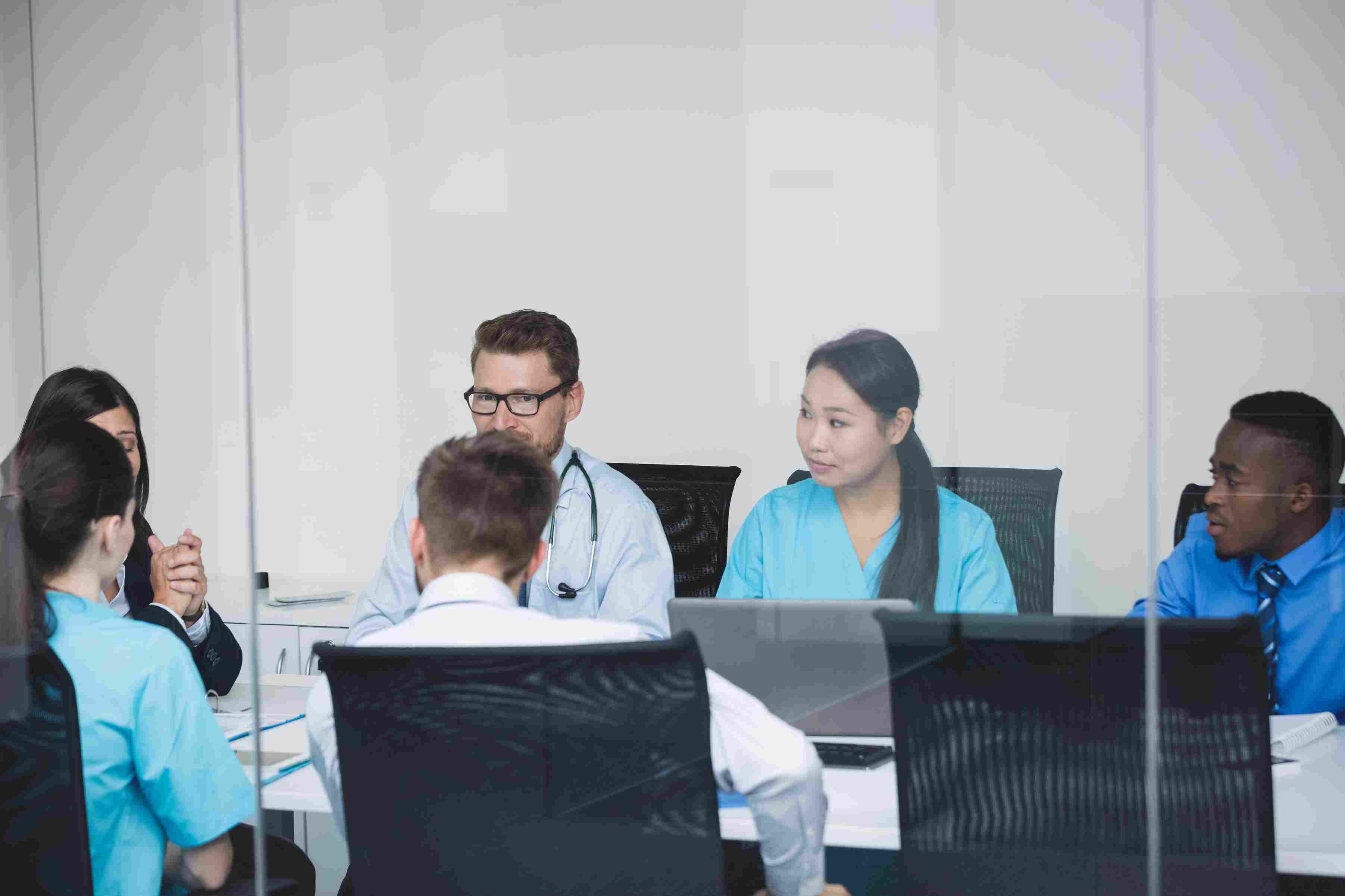 A diverse group of doctors, nurses, and hospital staff seated around a conference table in a glass-walled meeting room, representing the staff training and user acceptance testing phases of a Hospital Management System implementation