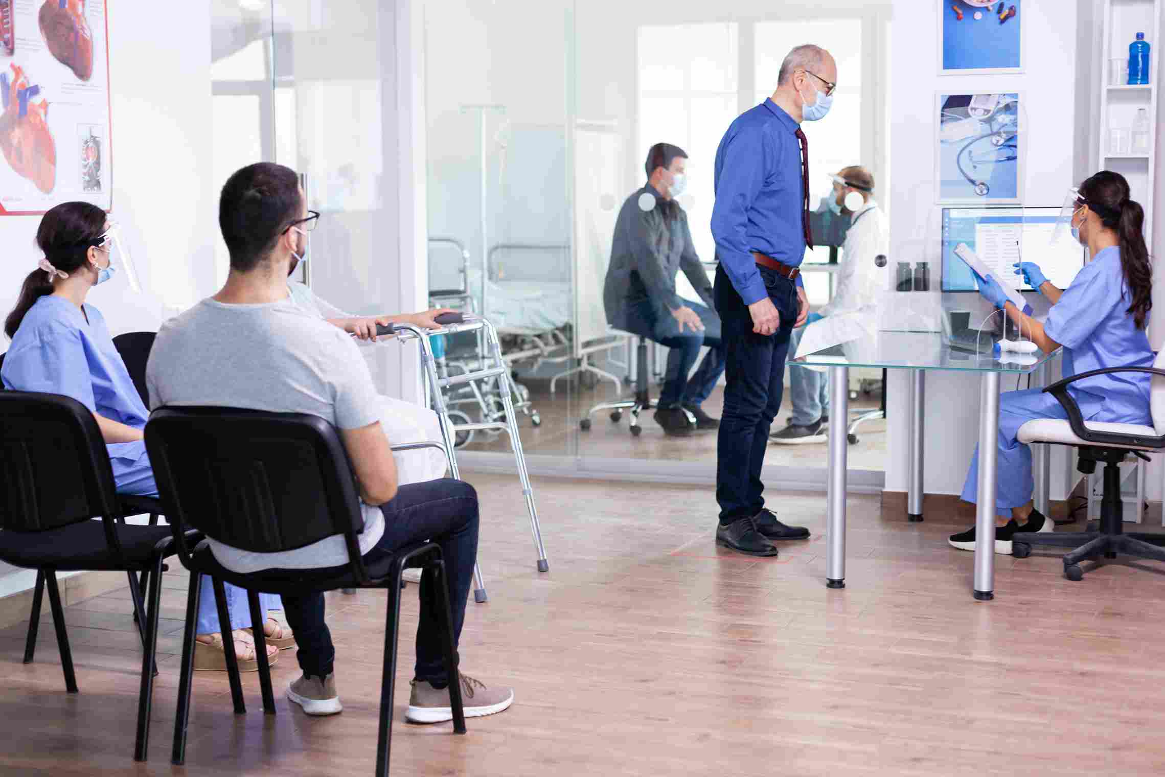 An elderly patient using a walking frame speaking with a nurse at a hospital reception desk while other patients wait, representing the patient registration and scheduling challenges that HMS implementation directly addresses
