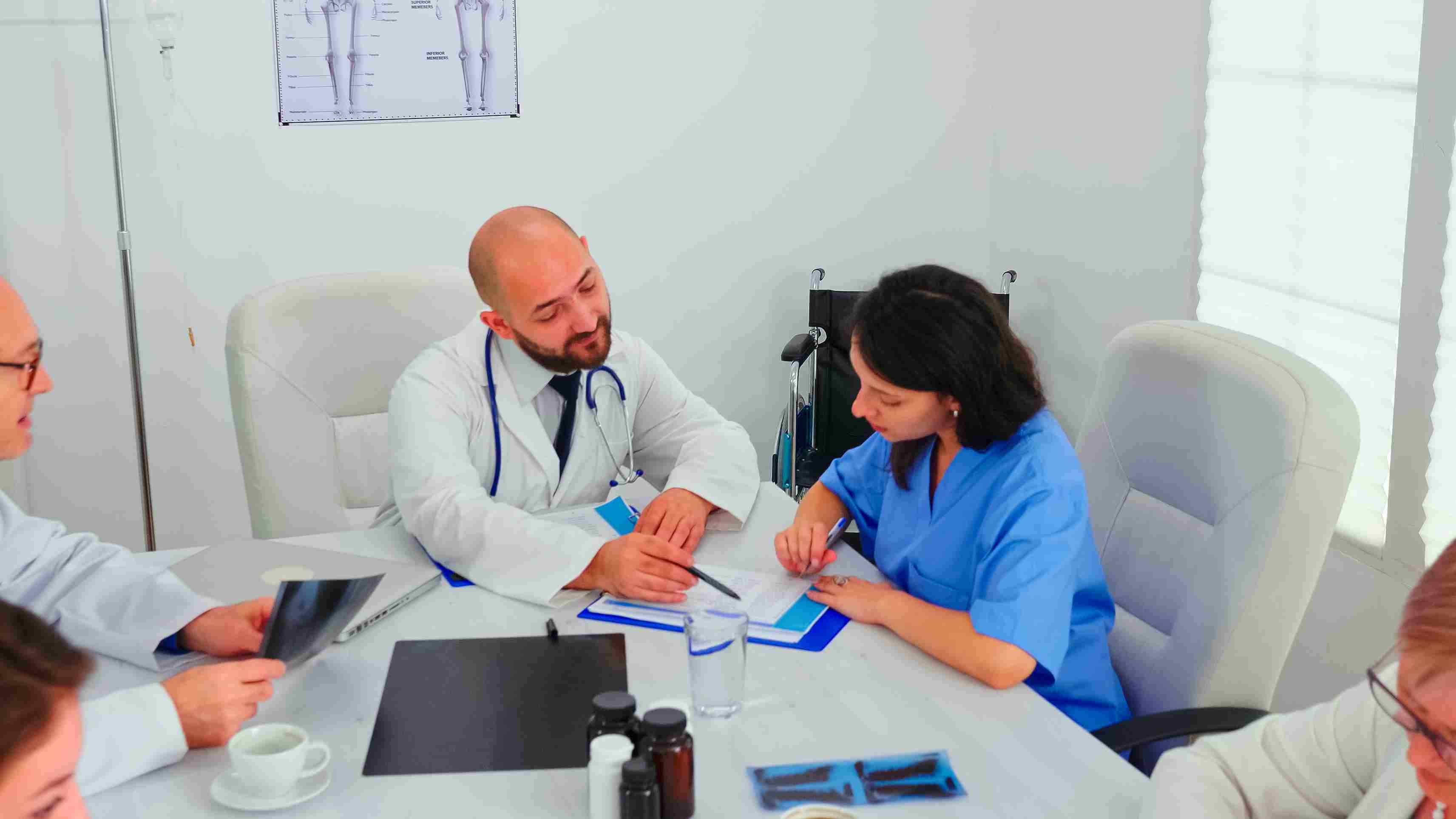 A senior doctor in a white coat explaining clinical documentation and compliance requirements to a diverse group of medical staff in a hospital conference room with radiography scans visible on screens in the background, representing the staff training, credentialing, and Human Resource Management chapter requirements of NABH 6th Edition accreditation that hospitals must fulfil through structured documentation supported by a Hospital Management System