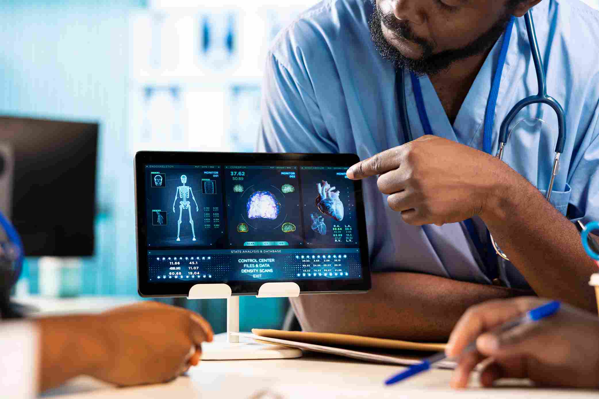 A doctor in blue scrubs pointing at a tablet displaying AI-powered clinical data including skeletal scans, brain imaging, cardiac data, and a stats analysis dashboard while a patient takes notes, representing the Embedded AI Mode of the ChatGPT Epic EHR integration where AI-assisted clinical data appears directly within the clinician's existing workflow during a patient consultation