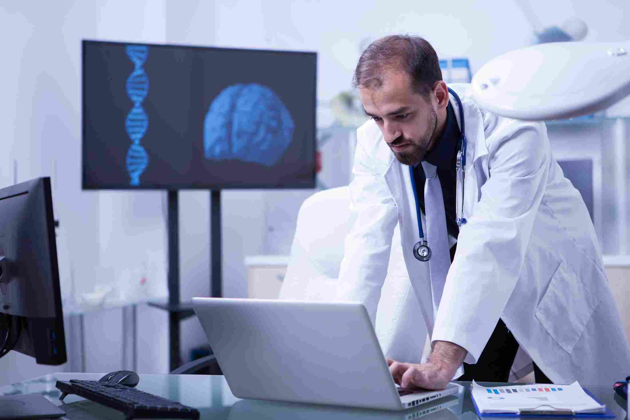 A focused doctor in a white coat leaning over a laptop in a clinical office with a digital brain scan and DNA model displayed on a monitor in the background, representing a clinician using the ChatGPT preparation tool mode to review patient records and identify clinical changes before walking into a consultation room