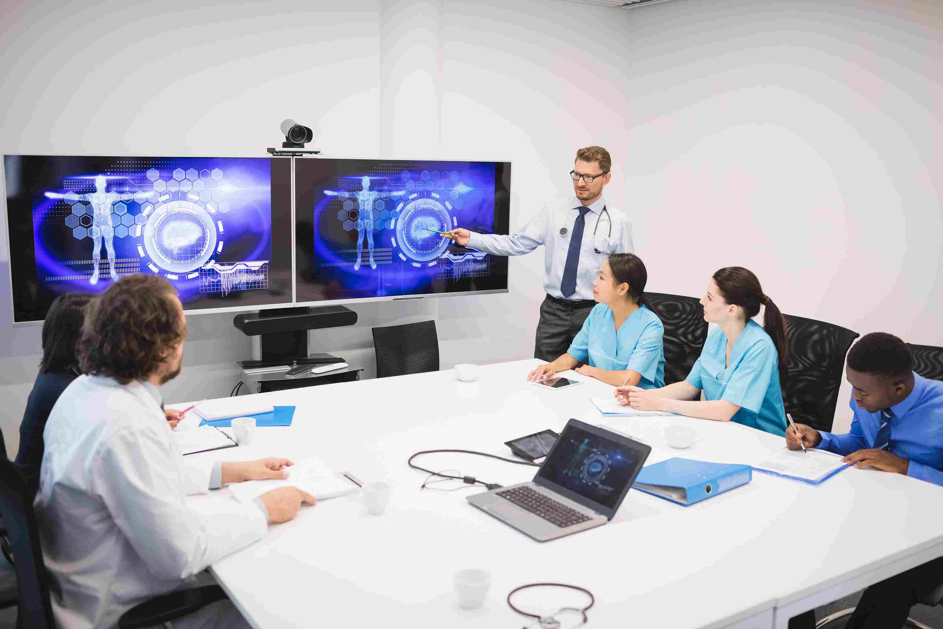 A doctor in a shirt and tie presenting AI-powered clinical data visualisations on a large digital screen to a team of nurses and medical staff seated around a conference table with laptops, representing a hospital CIO or clinical informatics team evaluating the two ChatGPT Epic EHR integration deployment modes before go-live